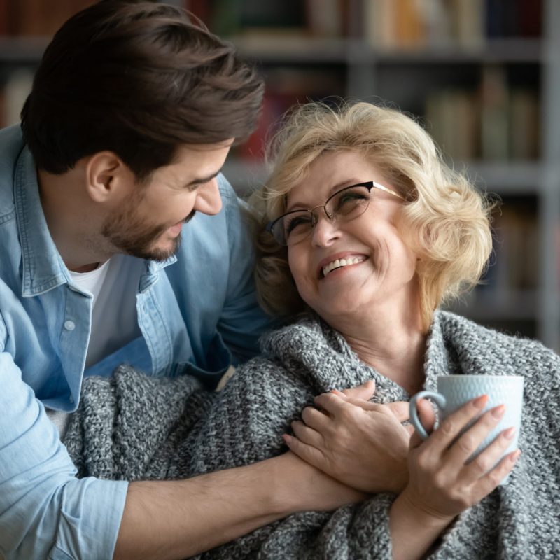 Caring young man covering happy middle aged old mother with plaid, giving cup of hot tea, showing love and devotion at home. Smiling retired woman enjoying peaceful comfort moment with grown son.
